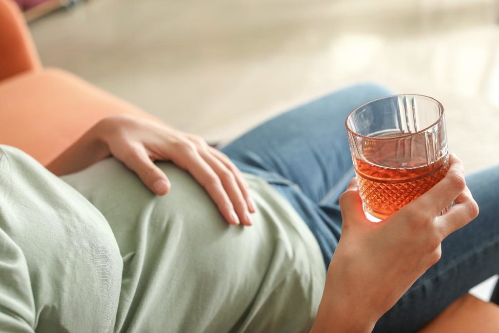 a woman touches her pregnant belly while holding onto an alcoholic drink and realizing there are many dangers of drinking during pregnancy