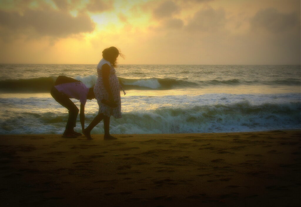 A pregnant woman and her husband walk the beach while talking